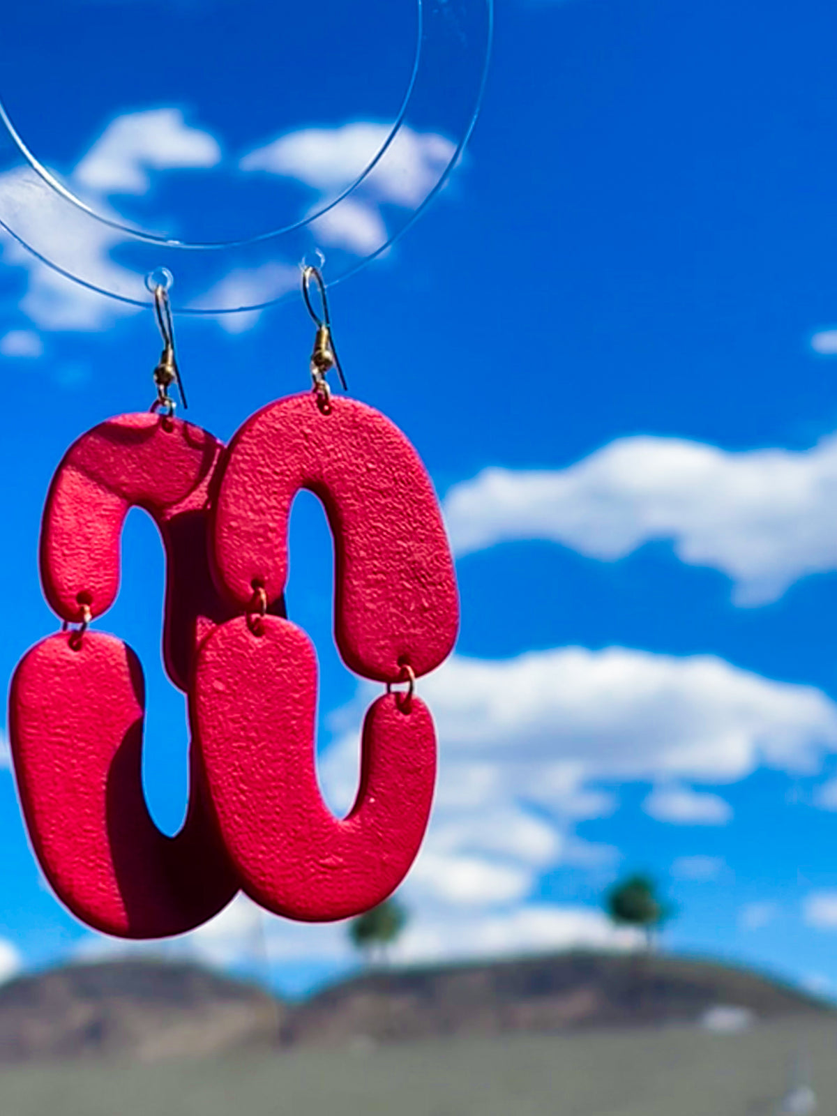 A pair of red, arc-shaped earrings hanging against a blue sky with clouds in the background.